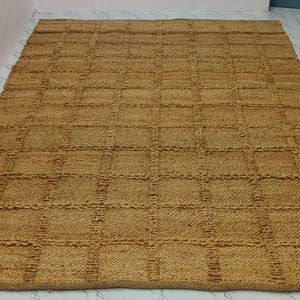 May include: A rectangular jute rug with a grid design. The rug is a warm, natural tan colour and appears to be made of thick, durable fibres. The rug is placed on a light-coloured floor.