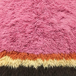 May include: A shaggy rug featuring a large section of pink fibers followed by horizontal bands of orange, cream, and dark brown. The long, dense pile creates a textured surface with distinct color transitions across the rectangular floor covering.