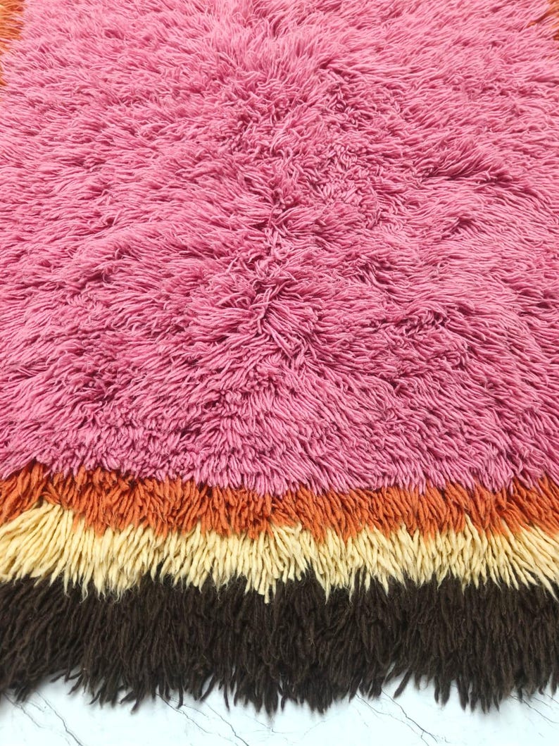 May include: A shaggy rug featuring a large section of pink fibers followed by horizontal bands of orange, cream, and dark brown. The long, dense pile creates a textured surface with distinct color transitions across the rectangular floor covering.