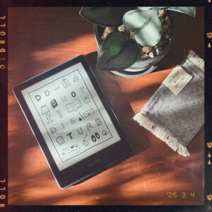 May include: A black Kindle e-reader with a white screen displaying the text "Do Not Disturb" with various icons. The e-reader is on a wooden surface with a gray and white fringed fabric with the text "Everyday with DePround" on it.