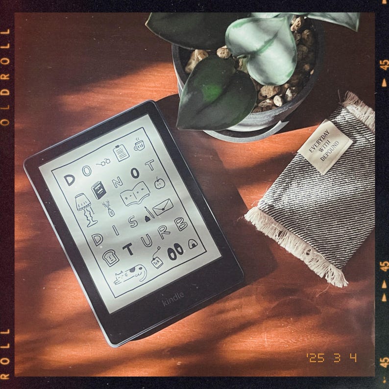 May include: A black Kindle e-reader with a white screen displaying the text "Do Not Disturb" with various icons. The e-reader is on a wooden surface with a gray and white fringed fabric with the text "Everyday with DePround" on it.