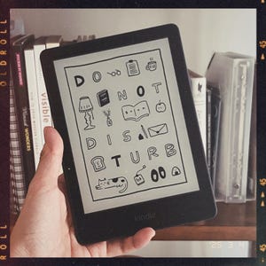 May include: A black and white Kindle e-reader displaying a digital illustration of the phrase "Do Not Disturb" with various icons and doodles. The Kindle is on a bookshelf with other books.