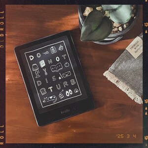 May include: A black and white Kindle e-reader with the text "DO NOT DISTURB" displayed on the screen. The text is surrounded by small icons, including a lamp, a book, a coffee cup, and a cat.