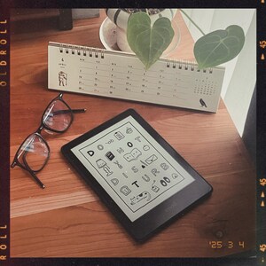 May include: A black e-reader with a white screen displaying a list of words and icons. The words include "Do Not Disturb". The e-reader is sitting on a wooden table with a calendar and a pair of glasses.