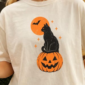 May include: An ivory short-sleeve t-shirt featuring a graphic print of a black cat sitting on top of an orange jack-o'-lantern. Above the cat is a large orange full moon with a small black bat silhouette and several small orange four-pointed stars.