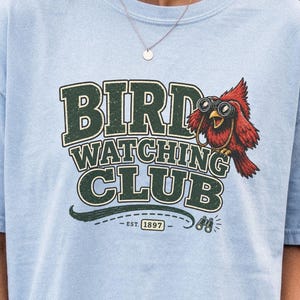 May include: A light blue t-shirt featuring a graphic with the text BIRD WATCHING CLUB and a red cardinal bird wearing binoculars. The design includes the text EST. 1897 and a small illustration of binoculars below the main lettering.