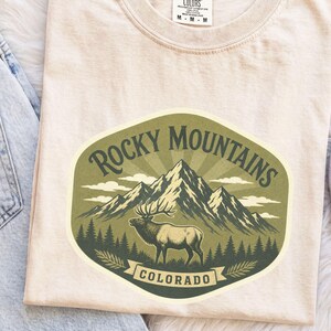 May include: An ivory t-shirt featuring a graphic of the Rocky Mountains in Colorado with an elk standing in front of a mountain range and pine trees. The design is enclosed in an oval shape with the text Rocky Mountains and Colorado.