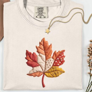 May include: An ivory t-shirt featuring a central graphic of a maple leaf composed of various fabric patterns including plaid, polka dots, and floral prints in shades of red, orange, and yellow. The shirt has a Comfort Colors brand label.