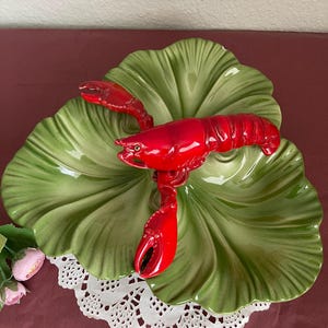 May include: A ceramic serving dish with a green, leaf-shaped design, topped with a bright red lobster figurine. The dish sits on a white lace doily, creating a coastal theme.