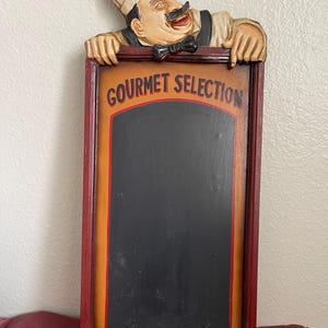 May include: A rectangular chalkboard sign with a smiling chef figure at the top. The sign has a black center with the words "GOURMET SELECTION" in red letters. The frame is red and orange.