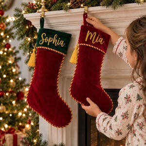 Op de afbeelding: Twee kerstsokken van fluweel hangen aan een schoorsteenmantel. De linker sok is donkerrood met een donkergroene boord waarop de naam Sophia in goud is geborduurd. De rechter sok is donkerrood met de naam Mia in goud geborduurd. Gouden afwerking.