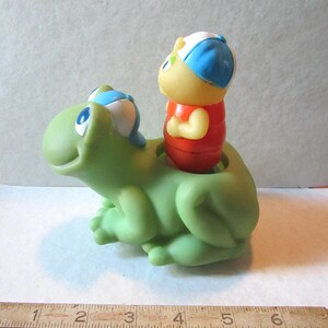 Puede incluir: Un juguete de pl&aacute;stico verde con forma de rana sonriente y una figura roja, amarilla y azul montada en su espalda. El juguete tiene una est&eacute;tica vintage y es probablemente un juguete para ni&ntilde;os.