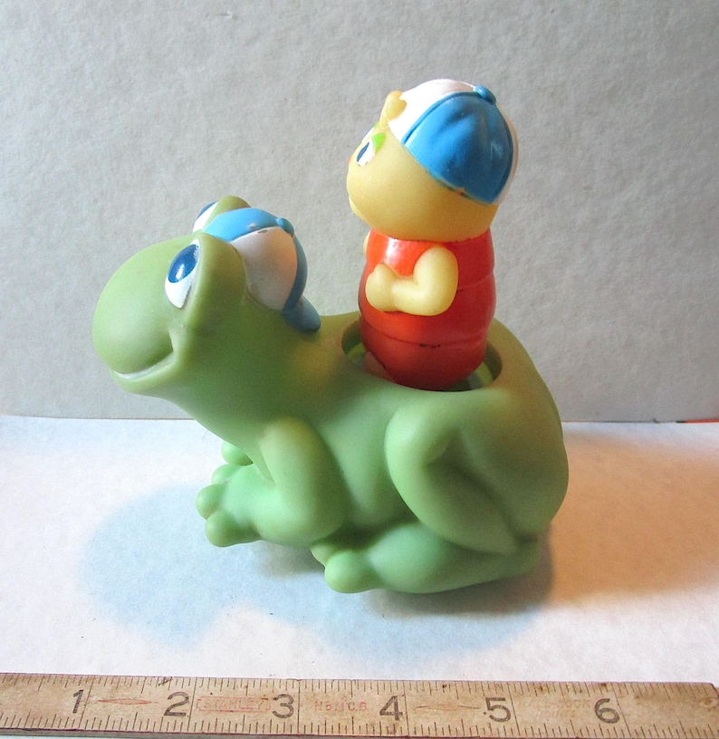Puede incluir: Un juguete de pl&aacute;stico verde con forma de rana sonriente y una figura roja, amarilla y azul montada en su espalda. El juguete tiene una est&eacute;tica vintage y es probablemente un juguete para ni&ntilde;os.