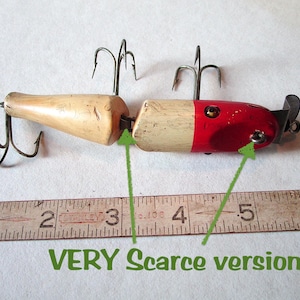 May include: Vintage fishing lure with a wooden body and metal hooks. The lure is painted red, white, and natural wood tones. The text "VERY Scarce version!" is visible. The lure is approximately 5 inches long.