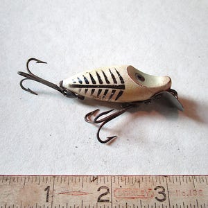 May include: Vintage fishing lure with a cream-colored body and black markings. The lure has two treble hooks and a small metal fin. A wooden ruler is in the foreground, showing measurements in inches.