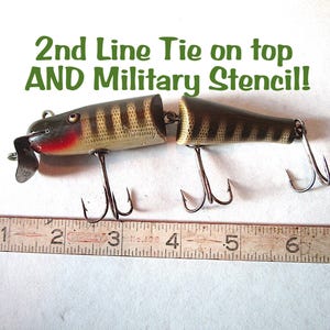 May include: Vintage fishing lure with a segmented body, featuring a brown and tan striped pattern. The lure has a red and black head, a metal lip, and multiple treble hooks. The text "2nd Line Tie on top AND Military Stencil!" is visible.