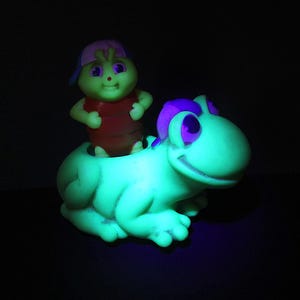 Puede incluir: Una rana de juguete verde con detalles morados y una cara sonriente. Una peque&ntilde;a figura verde con una camisa roja y una gorra rosa y azul est&aacute; sentada en la espalda de la rana. El juguete brilla en la oscuridad.