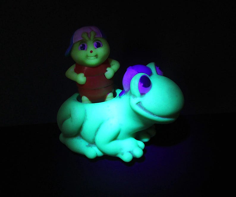 Puede incluir: Una rana de juguete verde con detalles morados y una cara sonriente. Una peque&ntilde;a figura verde con una camisa roja y una gorra rosa y azul est&aacute; sentada en la espalda de la rana. El juguete brilla en la oscuridad.