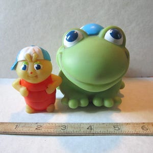 Puede incluir: Dos juguetes de pl&aacute;stico vintage: una figura amarilla y roja con una gorra azul y blanca, y una gran rana verde con grandes ojos azules y un sombrero azul. Una regla de madera est&aacute; en la parte inferior de la imagen.