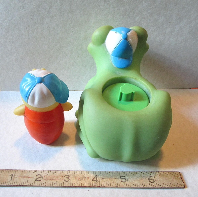 Puede incluir: Dos juguetes de pl&aacute;stico: un recipiente verde con forma de rana y una peque&ntilde;a figura naranja y amarilla. Ambos juguetes tienen gorras de b&eacute;isbol blancas y azules. La rana tiene un interior verde. Una regla de madera est&aacute; en la parte inferior de la imagen.