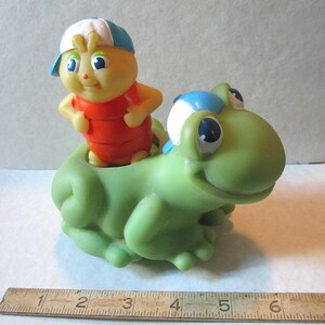 Puede incluir: Un juguete vintage que presenta una figura amarilla con forma de insecto con un atuendo rojo y una gorra azul y blanca, montada en una rana verde. El juguete tiene ojos azules grandes y es de pl&aacute;stico.