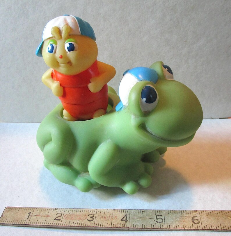 Puede incluir: Un juguete vintage que presenta una figura amarilla con forma de insecto con un atuendo rojo y una gorra azul y blanca, montada en una rana verde. El juguete tiene ojos azules grandes y es de pl&aacute;stico.