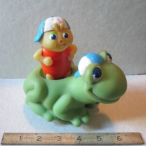 Puede incluir: Un juguete vintage que presenta una figura amarilla con forma de insecto con un atuendo rojo y una gorra azul y blanca, montada en una rana verde. El juguete tiene ojos azules grandes y est&aacute; hecho de un material pl&aacute;stico blando. Una regla de madera en la parte inferior.