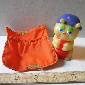 May include: A yellow glow-in-the-dark worm toy with a blue hat and green eyes. The worm is wearing a red and white outfit and is sitting on a bright orange fabric pouch.