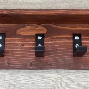 May include: A wooden wall-mounted shelf with five black metal hooks. The shelf is stained a dark brown color and has a rustic appearance.