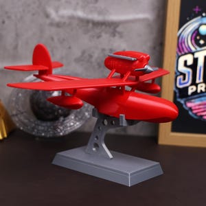 以下が含まれることがあります： グレーのスタンドに置かれた赤い飛行機の模型。飛行機はプロペラとフロートを備えています。飛行機は長方形のベースを持つグレーのスタンドの上にあります。