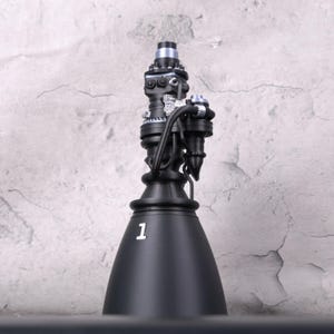 May include: A black model of a rocket engine, featuring intricate details and metallic accents. The base is cone-shaped with the number "1" in white. The top section has silver and black components, with tubes and nozzles.