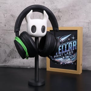 May include: Black gaming headphones with green cable resting on a black headphone stand. The stand features a white, cartoon-like character head. A framed print with the text "ELLAR PRINT LABS" is in the background.
