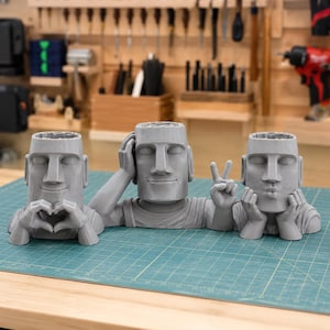 May include: Three gray 3D-printed Moai head planters arranged in a row. The left planter forms a heart shape with its hands, the center planter rests its head on one hand while making a peace sign, and the right planter rests its chin on both hands.