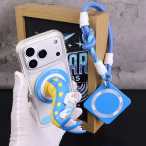 Könnte beinhalten: Transparente Handyhülle mit einem blau-gelben Gyarados-Design. Die Hülle hat einen blauen Ring mit dem Text "GYARADOS" und ein blaues Schlüsselband mit einem blauen quadratischen Anhänger. Die Handyhülle wird von einer weißen behandschuhten Hand gehalten.