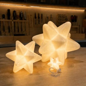 May include: Three multi-pointed star-shaped lamps in varying sizes, each emitting a warm, soft glow. The lamps feature a textured, off-white surface with geometric facets. A small clear plastic cord with a switch is visible in the foreground.