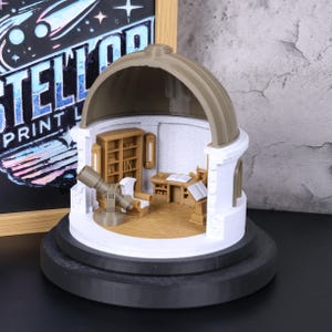 Op de afbeelding: Een miniatuur observatoriummodel met een koepeldak, met een telescoop, bureau en boekenplanken. Het model is voornamelijk wit en bruin, met een zwarte basis. De achtergrond bevat een schoolbord met de tekst "STELLAR PRINT LAB".
