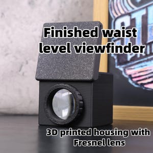 Puede incluir: Un visor de nivel de cintura negro impreso en 3D con una lente de Fresnel. El visor tiene una parte superior gris oscuro texturizada y una lente circular. El texto "Finished waist level viewfinder" y "3D printed housing with Fresnel lens" son visibles.