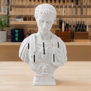 May include: A light gray stone-textured bust of a Roman figure wearing a toga and armor. The chest plate features a central Gorgon head relief and an eagle emblem at the base. Four vertical rectangular slots are cut into the front of the torso.