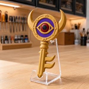 May include: A gold-colored key-shaped object featuring a circular head with two upward-curving horns and a central purple disc containing a red and black eye design. The object is mounted on a clear acrylic stand, showcasing its detailed relief.