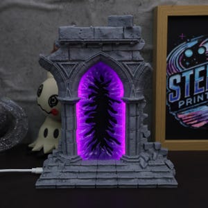 Può includere: Un arco grigio, simile alla pietra, con un portale viola brillante. L'arco ha un design dettagliato e strutturato ed è illuminato dall'interno. Un cavo USB bianco è collegato alla base. Una stampa incorniciata con il testo "STE PRINT" è sullo sfondo.
