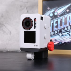 May include: A white action camera with a black lens and a red accent ring. The camera has a black screen and a red dial. The word "ACTION 5" is printed on the side. It is mounted on a black base with a white adjustment knob.
