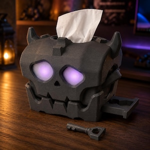 May include: A dark gray, skull-shaped tissue box holder with two glowing purple eyes and small horns. The box features a top opening for tissues and a small side drawer. A matching dark gray key is positioned in front of the skull-shaped container.