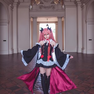 May include: A cosplayer in a black and red gothic-style dress with long sleeves and a flowing cape. The dress features a high collar, red accents, and a short skirt. The cosplayer has pink hair and is wearing black boots. The background includes a building with columns and a clock.