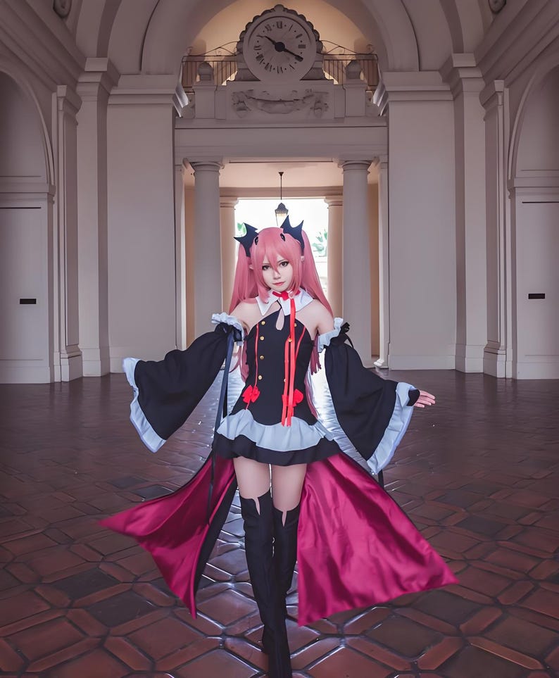 May include: A cosplayer in a black and red gothic-style dress with long sleeves and a flowing cape. The dress features a high collar, red accents, and a short skirt. The cosplayer has pink hair and is wearing black boots. The background includes a building with columns and a clock.