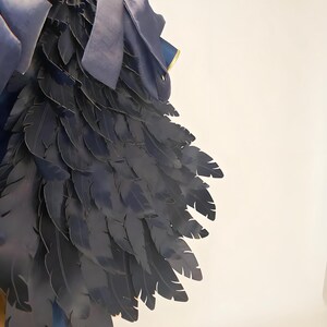 May include: Close-up of a dark blue costume piece with layered feather-like details. The design includes a satin bow and trim, suggesting a theatrical or fantasy theme. The feathers are arranged to create a wing-like shape.