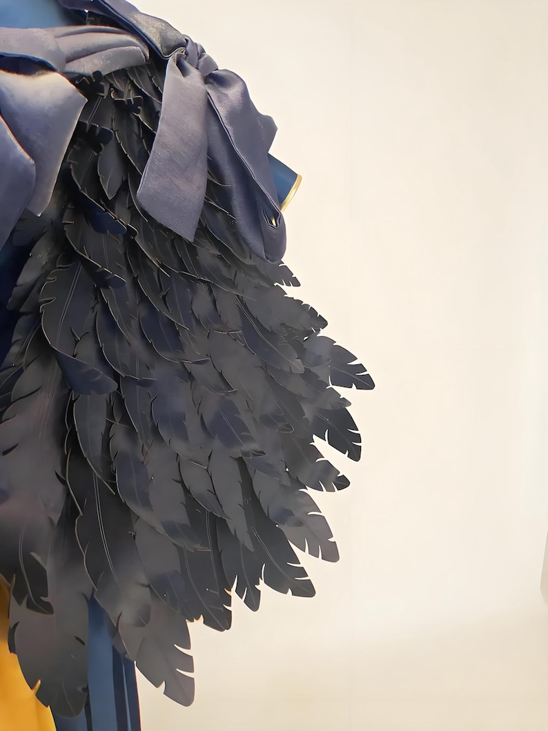 May include: Close-up of a dark blue costume piece with layered feather-like details. The design includes a satin bow and trim, suggesting a theatrical or fantasy theme. The feathers are arranged to create a wing-like shape.