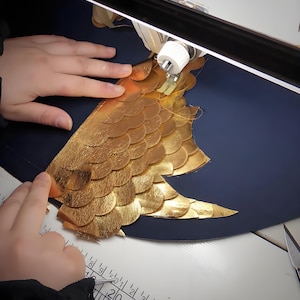 May include: A close-up of a sewing project in progress. Gold-colored, scale-like leather pieces are being sewn onto a dark blue surface. The project is illuminated by a bright light, with hands guiding the material.