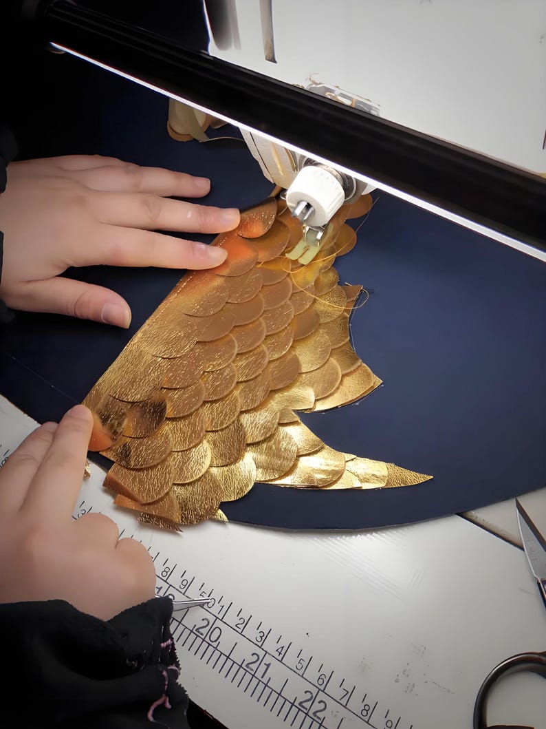 May include: A close-up of a sewing project in progress. Gold-colored, scale-like leather pieces are being sewn onto a dark blue surface. The project is illuminated by a bright light, with hands guiding the material.