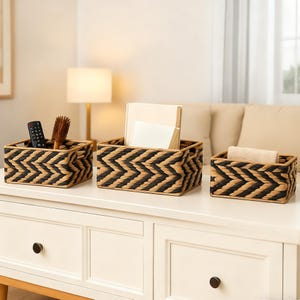 May include: Three rectangular woven storage baskets with a black and tan chevron pattern. The baskets are on a white surface. One basket holds a remote and brush, another holds papers, and the third holds a rolled towel.