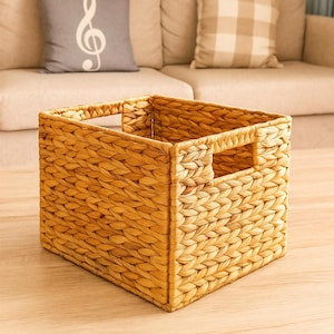 May include: A square woven storage basket with cut-out handles. The basket is made of a light brown, natural material and has a textured surface. The basket is empty and sits on a light-colored wooden floor.
